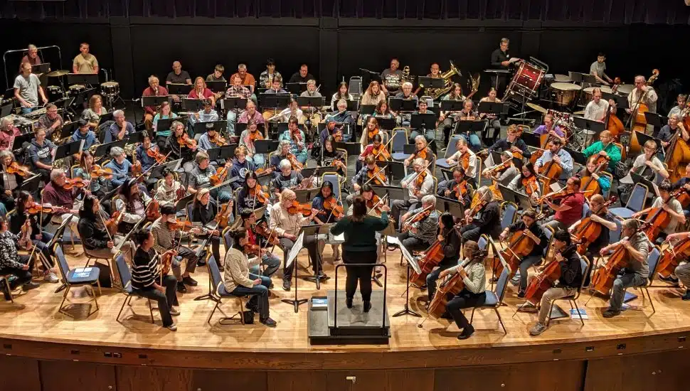 A large symphony orchestra rehearses on stage, with musicians playing strings, brass, woodwinds, and percussion, while a conductor leads from the podium in front.