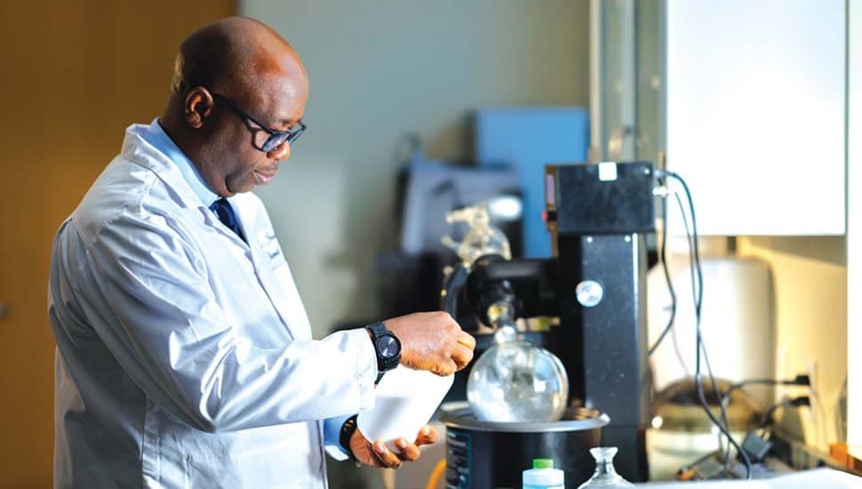 A chemist (Dr. Esosa Iriowen) in a lab coat carefully pours a substance into a beaker while working with laboratory equipment in a science lab.
