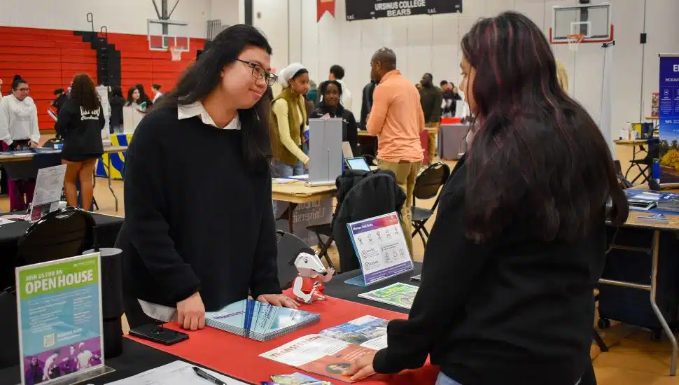 A student speaks with a college representative at a table during the Chester County Futures College Expo at Ursinus College, surrounded by other students and school booths in the gymnasium.