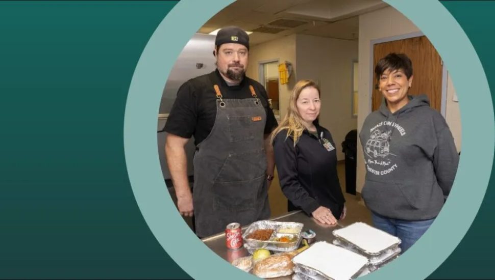 Three people stand together in a kitchen preparing meal trays, including a chef in an apron, a staff member in a uniform, and a volunteer wearing a Meals on Wheels sweatshirt. Packaged meals and ingredients are visible on the counter in front of them.