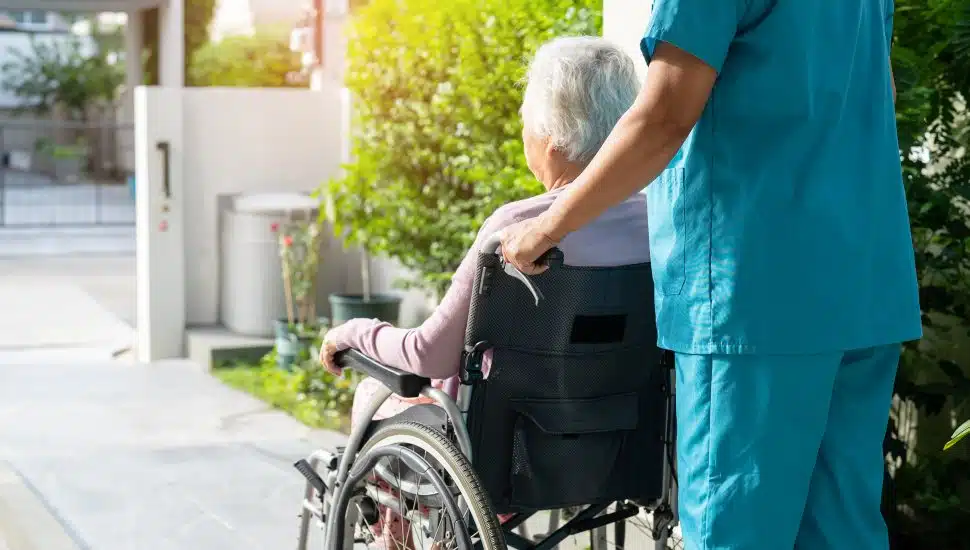 Nurse pushes elderly person in a wheelchair outside.