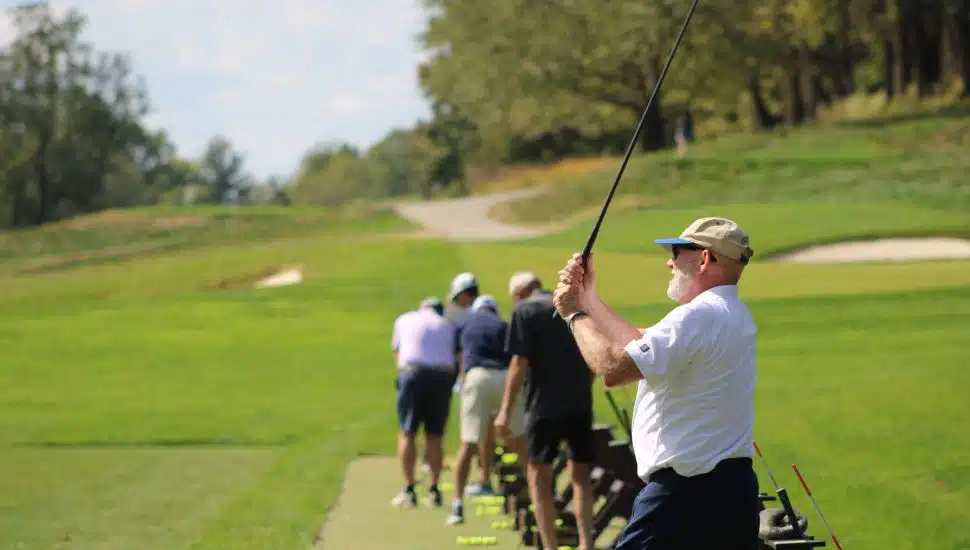 A line of golfers practicing their swings on a sunny day at a golf course during the Hankin Invitational charity tournament.