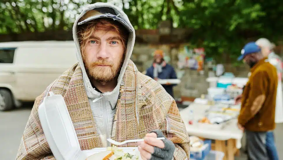 A bearded homeless man in warm clothing eating from a bowl at a charity event.