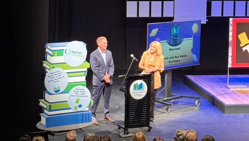 Sue and Frank Mattei stand at a podium speaking on stage during the Laughs for Literacy fundraiser, with a display of books and event signage behind them.