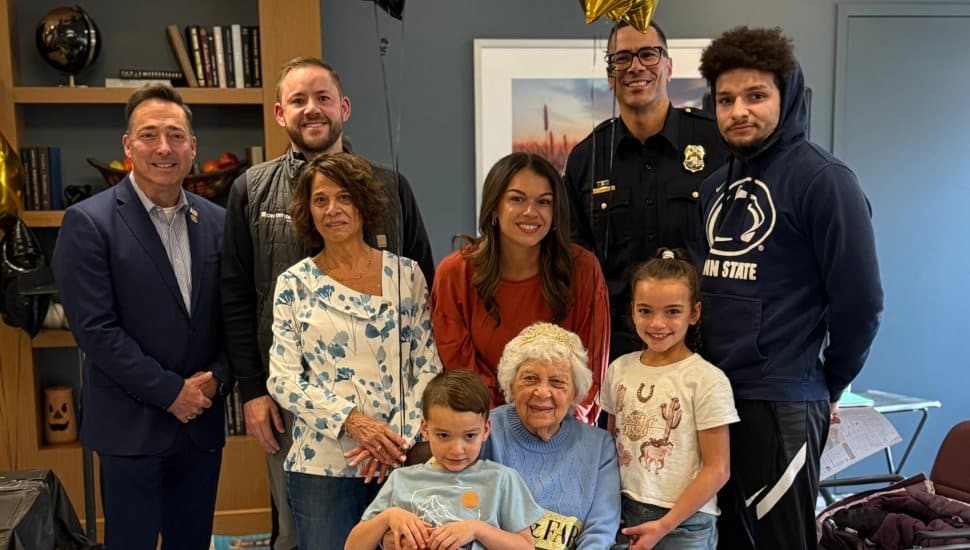 Angela “Lil” Laperuta, seated and smiling with a birthday crown, poses with family members, a police chief, and community leaders during her 100th-birthday celebration at Barclay Friends.
