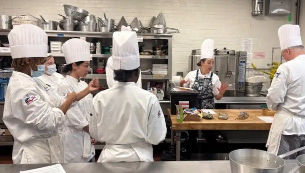 Culinary students in chef coats and hats gather around a workstation as a chef instructor demonstrates techniques in a commercial teaching kitchen.
