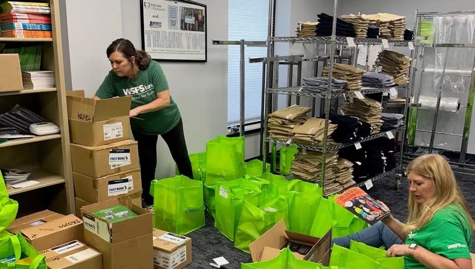 Two WSFS Bank volunteers sort donated books and supplies into bright green bags inside a Partners in Outreach workspace, surrounded by shelves of neatly organized clothing and boxes of materials.