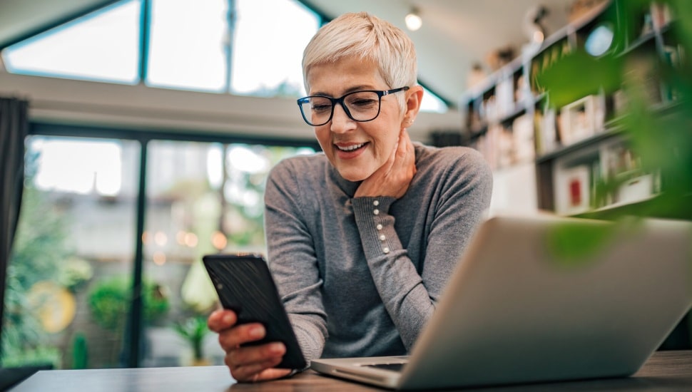 A senior woman sits at home looking at her laptop and smart phone.