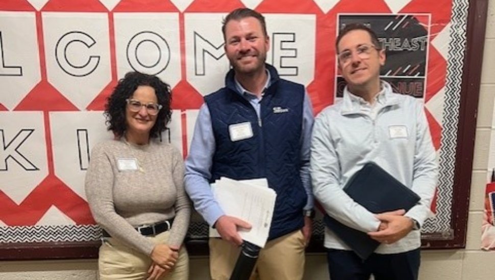 Three professionals from a community banking organization and a nonprofit stand indoors holding documents, smiling after a financial literacy workshop for students.