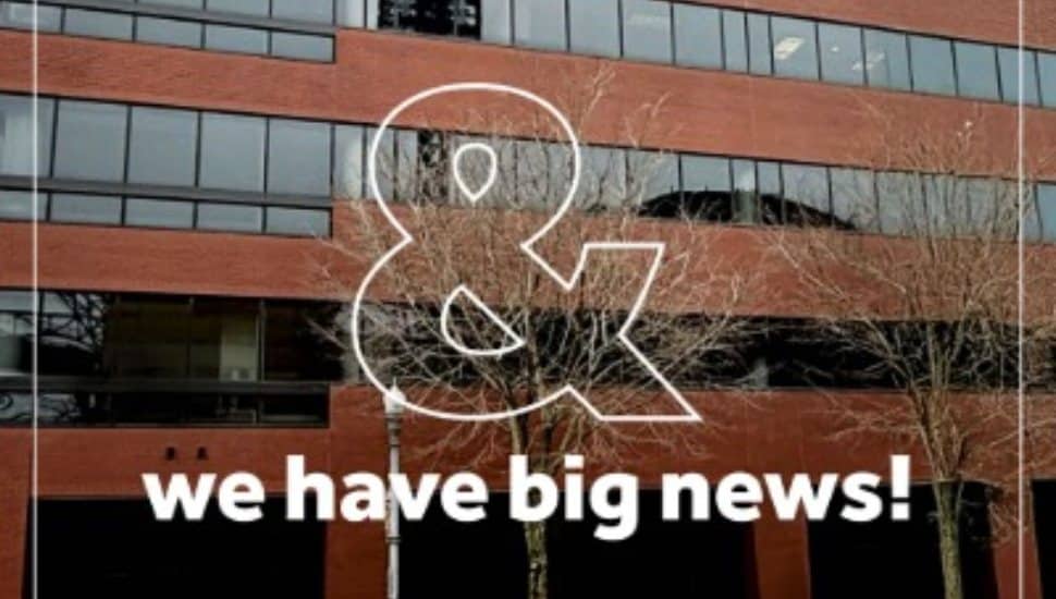Exterior of a multi-story office building with windows and trees in front, featuring an overlay graphic that reads “& we have big news!”