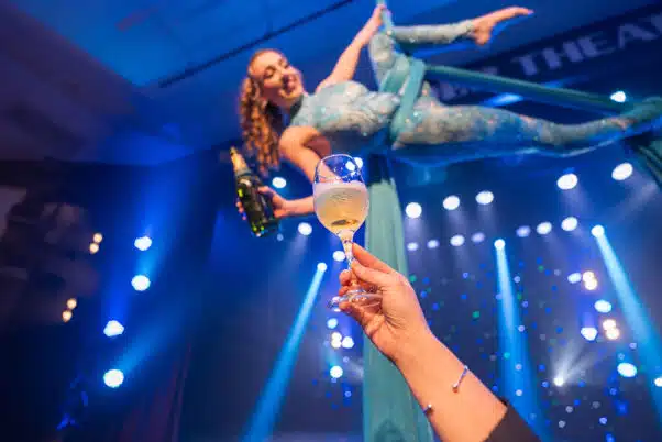 Aerial performer suspended in teal silks serving champagne to a guest during a dinner theater show under blue stage lighting.