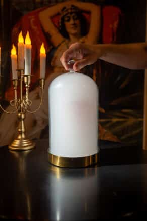 Hand lifting a glass cloche to reveal a smoke-filled cocktail presentation beside candlelight in a theatrical dining setting.