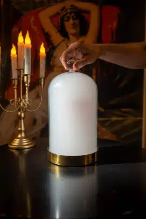 Hand lifting a glass cloche to reveal a smoke-filled cocktail presentation beside candlelight in a theatrical dining setting.