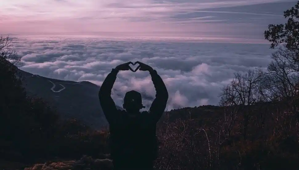 Person atop mountain makes a heart symbol with hands.