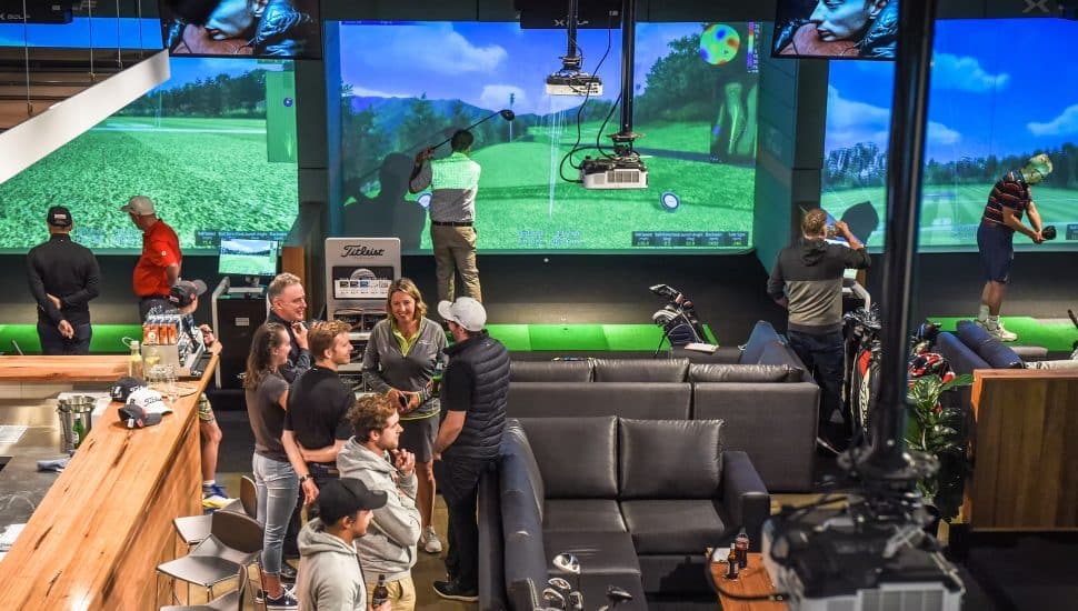 The inside of an X-Golf center highlighting golf simulators and people enjoying food and drink.