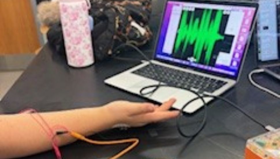 A student sits at a lab table with electrodes attached to their arm, connected by orange wires to a data collection device and laptop. The laptop screen displays a live green waveform graph, likely capturing muscle activity or biofeedback data. Lab equipment and notebooks are visible in the background, reinforcing the classroom laboratory setting.