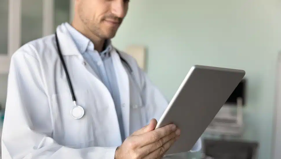 Male physician doctor in white coat using modern technology, working with tablet in hospital office, reading patients history records, medical examination electronic reports.