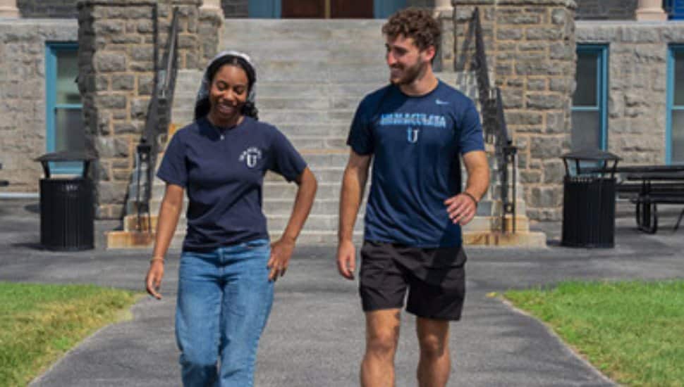 Two Immaculata University students wearing Immaculata T-shirts walk together on campus in front of a stone academic building.