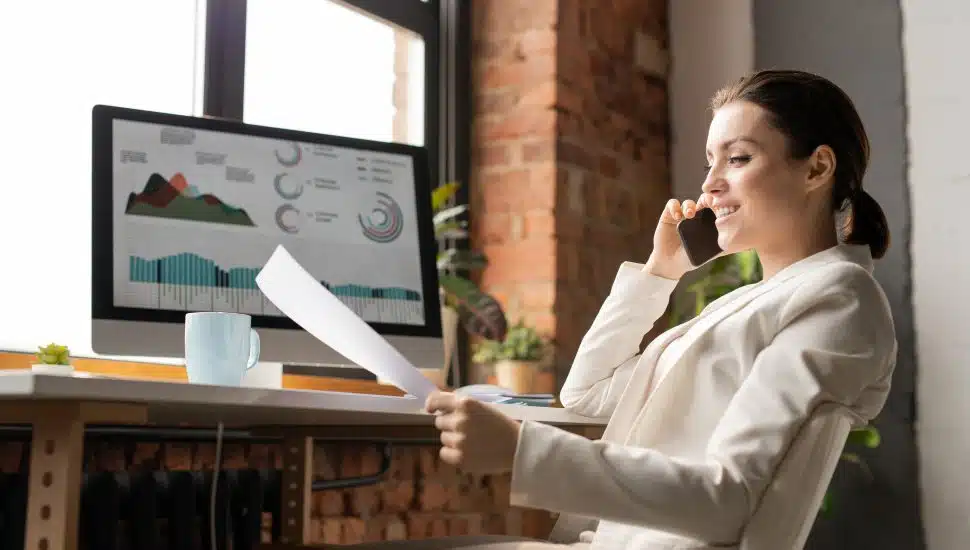Woman investor on phone call at desk while looking at paper.