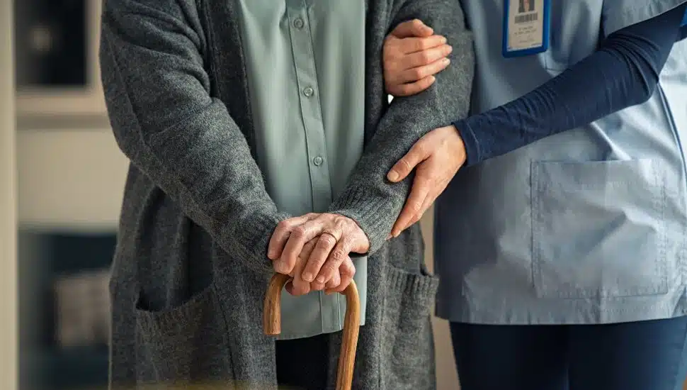 Nurse aide helps elderly person walk with cane.