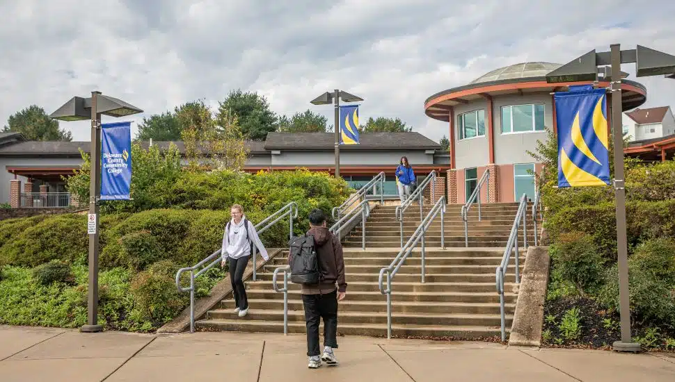 Students walk up and down the steps outside Delaware County Community College’s Downingtown Center, with campus banners and landscaping visible near the entrance.