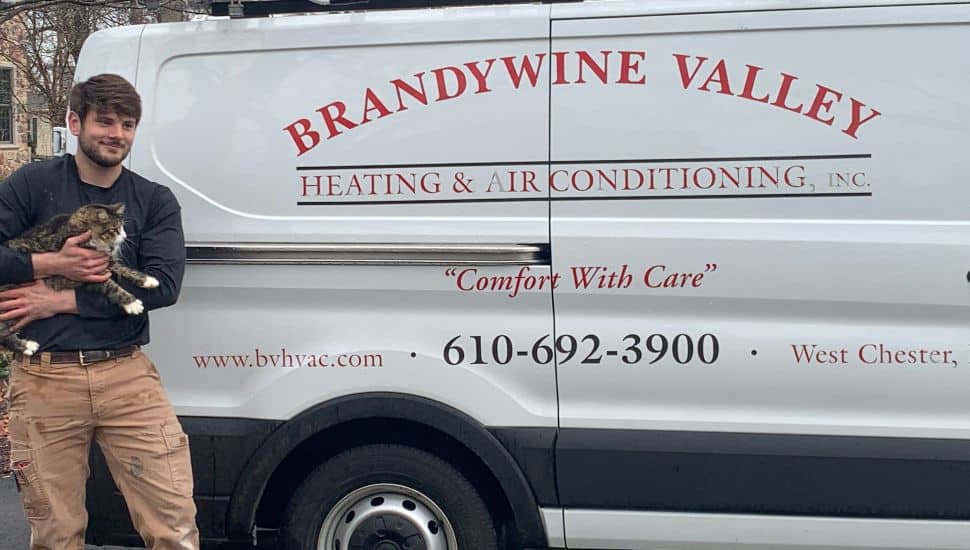 A Brandywine Valley Heating & Air Conditioning technician stands beside a company service van while holding a rescued tabby cat. The van displays the company’s name, website, phone number, and the slogan “Comfort With Care.” The scene appears to take place outdoors in a residential neighborhood.