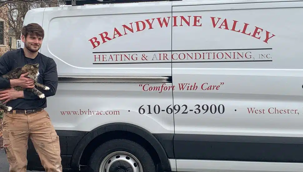 A Brandywine Valley Heating & Air Conditioning technician stands beside a company service van while holding a rescued tabby cat. The van displays the company’s name, website, phone number, and the slogan “Comfort With Care.” The scene appears to take place outdoors in a residential neighborhood.