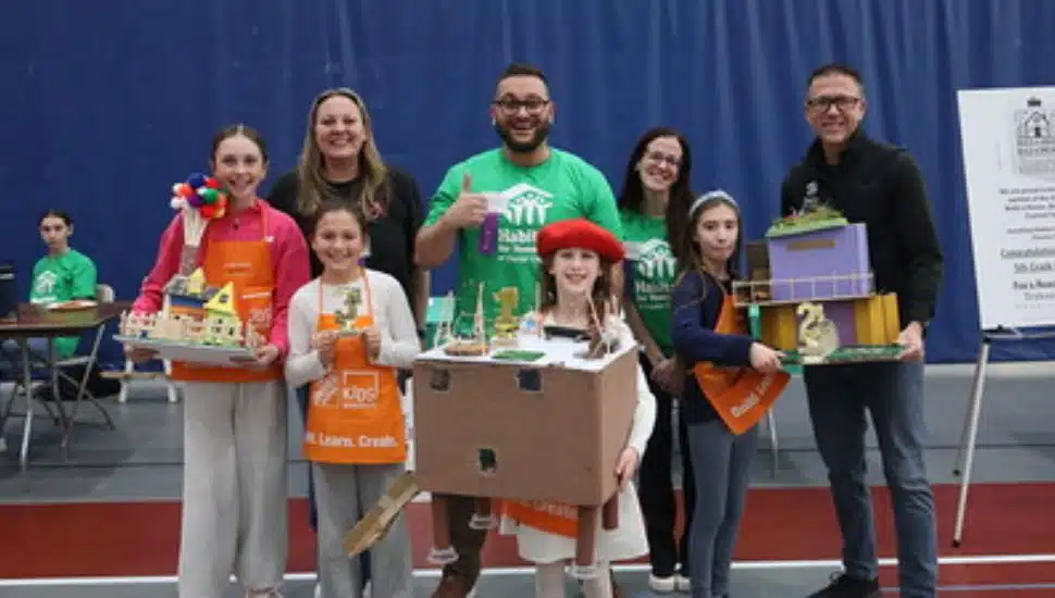 Group of elementary school students and adult volunteers standing in a gymnasium, smiling and holding colorful handmade model houses during a Habitat for Humanity student design competition event.