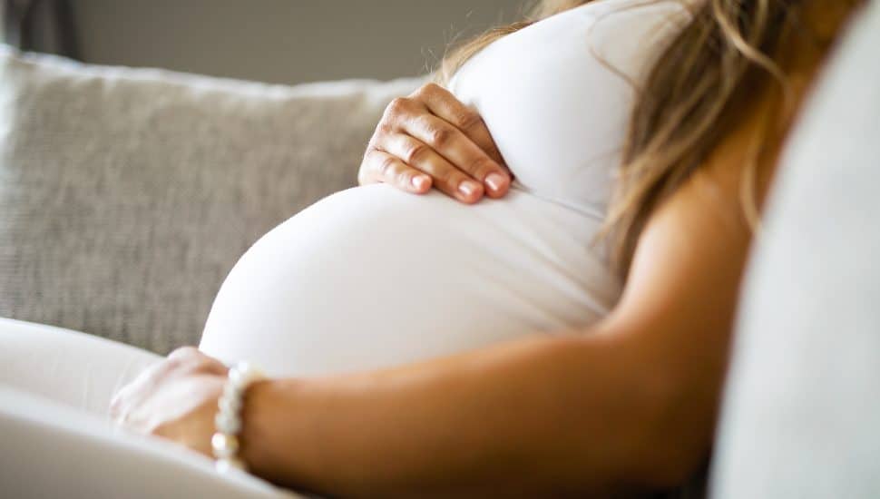 Close up of pregnant woman holding belly while sitting on couch.