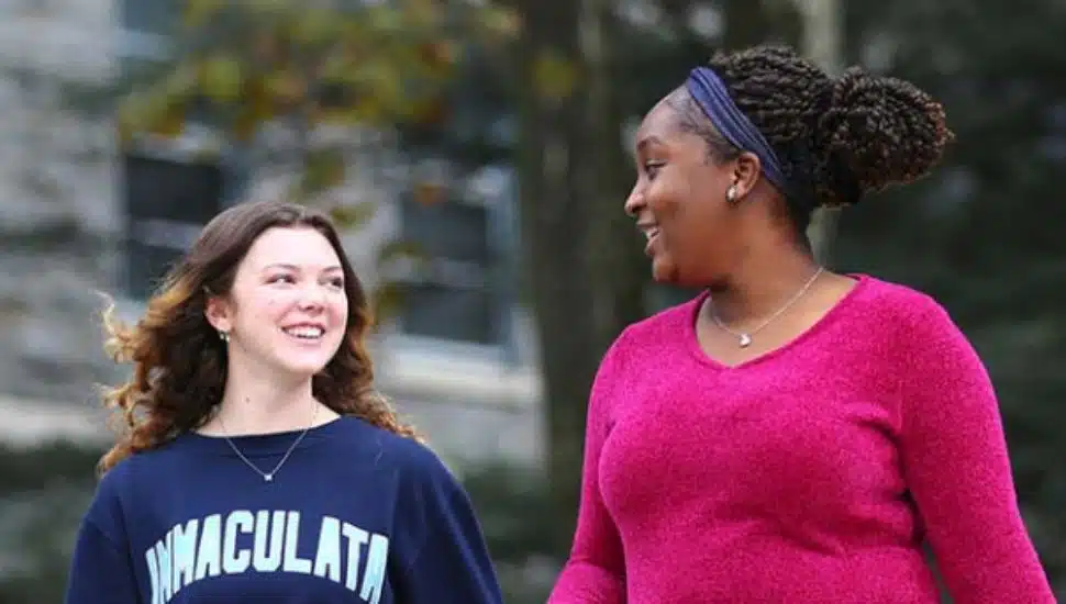 Two students walk side by side on a college campus pathway, smiling and chatting. One student wears a navy sweatshirt, while the other wears a bright pink sweater and has her hair pulled back with a headband.