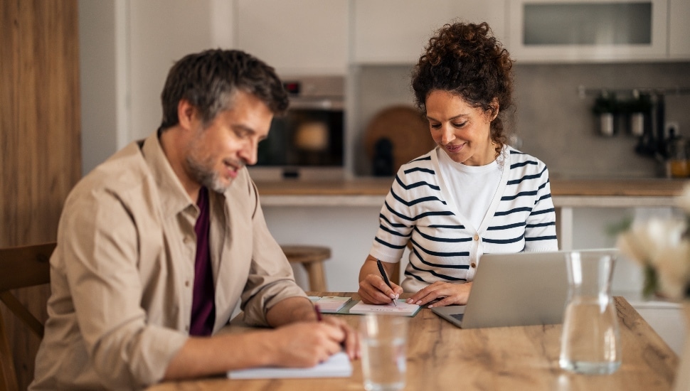 A husband and wife work together at a wooden kitchen table.