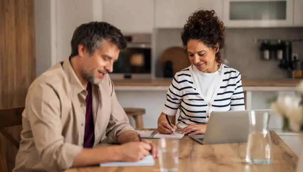 A husband and wife work together at a wooden kitchen table.