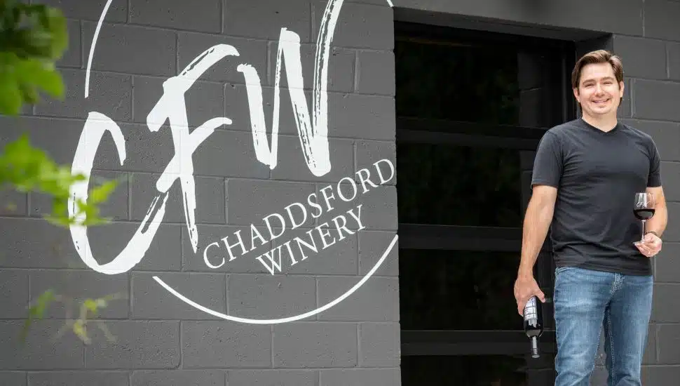 Corey Krejcik holding wine glasses and bottle next Chaddsford Winery signage.