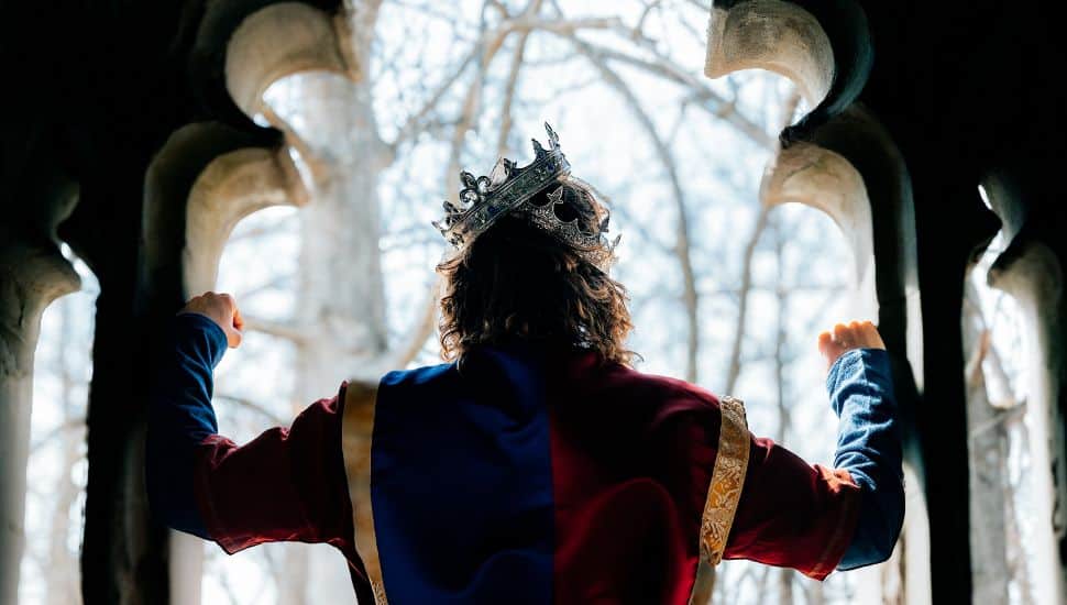 Actor portraying King Henry V wearing a crown and raising his arms triumphantly in a dramatic stage setting framed by gothic arches.