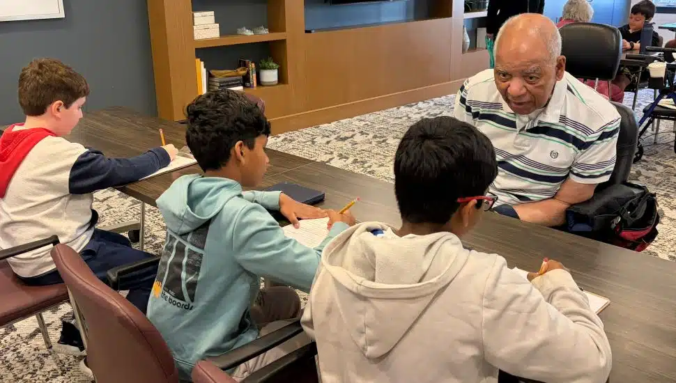 Three elementary school students sit at a table with an older adult resident, taking notes while he shares his experiences in a warm, community setting.