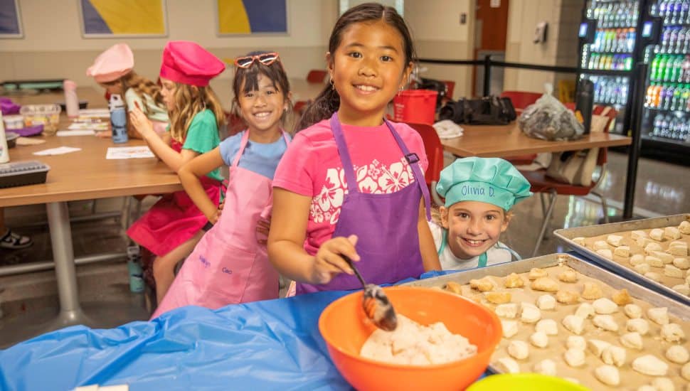 Children wearing aprons and chef hats participate in a cooking activity at Delaware County Community College Summer Kids Camps, preparing dough on baking trays in a classroom setting.
