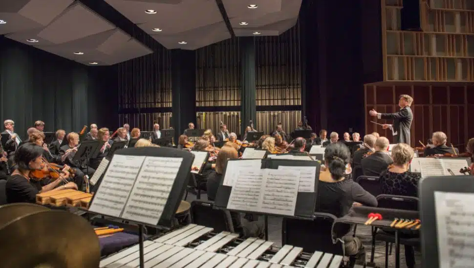 Orchestra performing on stage at Longwood Gardens, with a conductor leading musicians and visible sheet music in the foreground during a symphony concert.