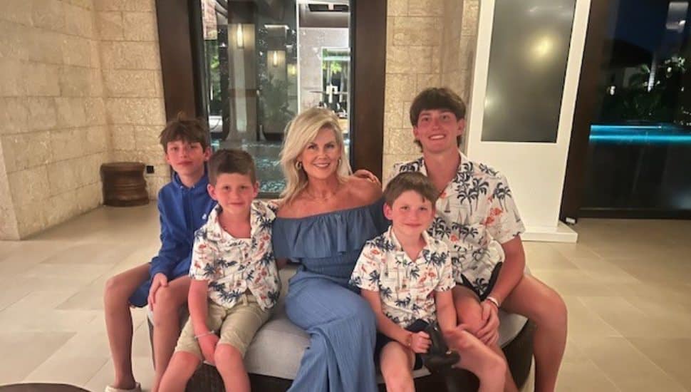 A woman in a blue dress sits on a bench with four boys around her, smiling together indoors.