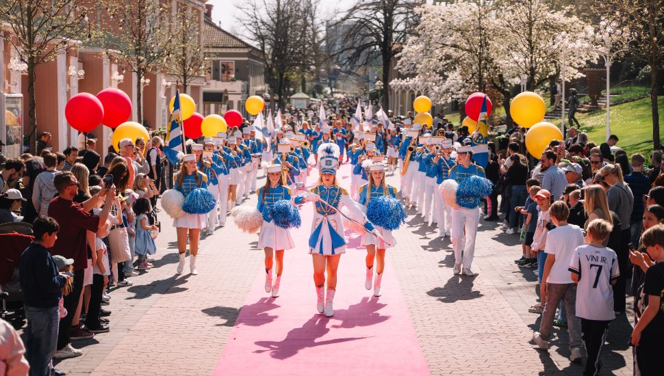 International marching band performers parade through a crowded street with colorful balloons and spectators during a WAMSB championship event tied to America’s 250th anniversary celebration.