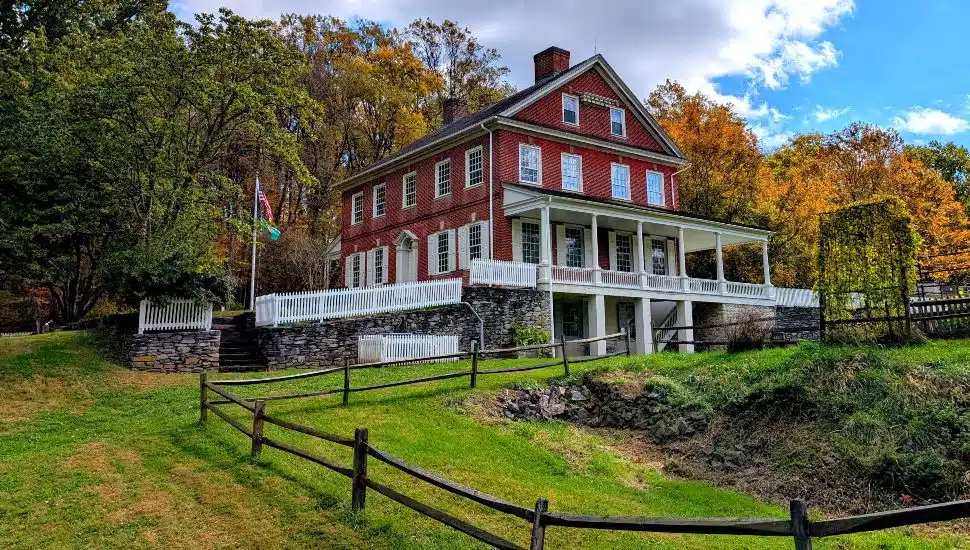 Historic Rock Ford exterior view surrounded by autumn trees, featuring the historic red brick mansion, wraparound porch, stone retaining walls, and sloping grassy landscape.