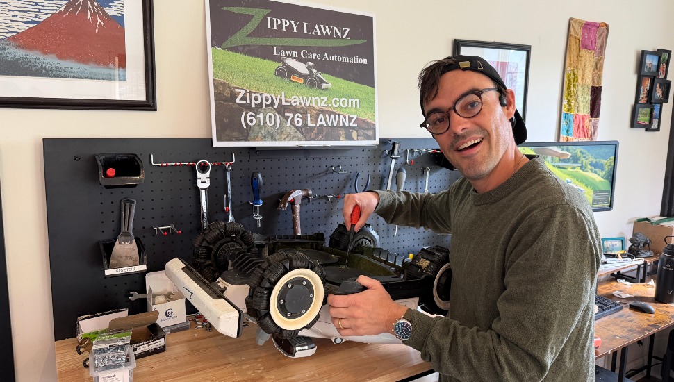 Paul Richards smiles while working on a disassembled robotic lawn mower at a pegboard workbench in his West Chester garage, with a Zippy Lawnz Lawn Care Automation sign visible on the wall behind him.