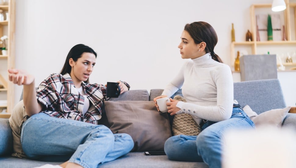 Two women sit on a couch. One speaks as the other listens.