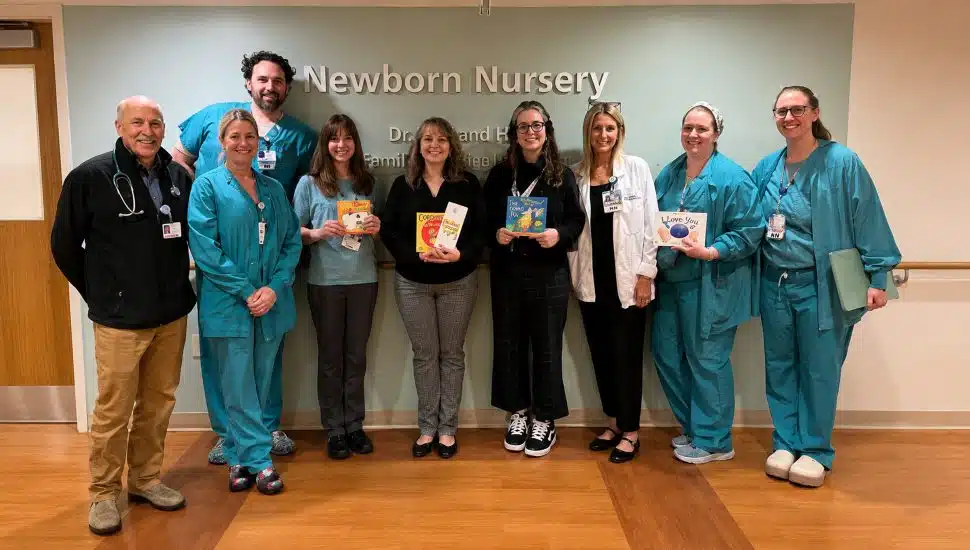 A group of nine people — several in teal scrubs — stand in front of a Newborn Nursery sign at Paoli Hospital, holding colorful children's board books donated by Easttown Library.