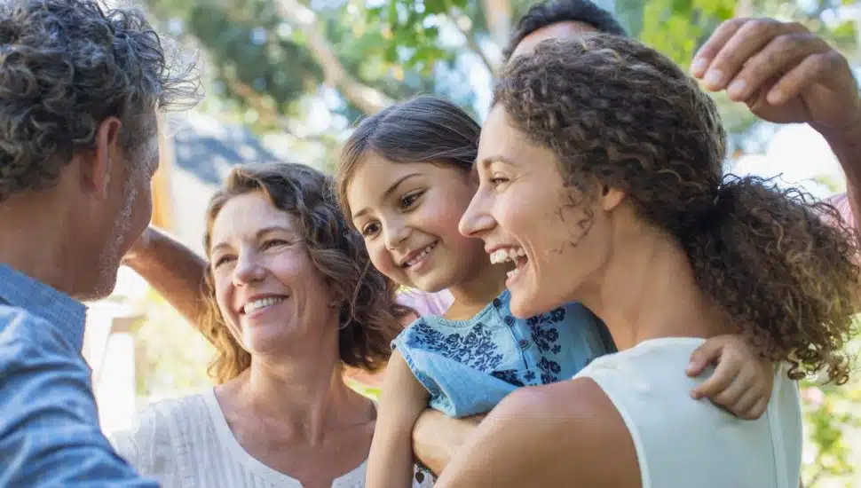 A smiling multigenerational family outdoors, with a young child being held and interacting warmly with adults in a joyful, sunlit setting.