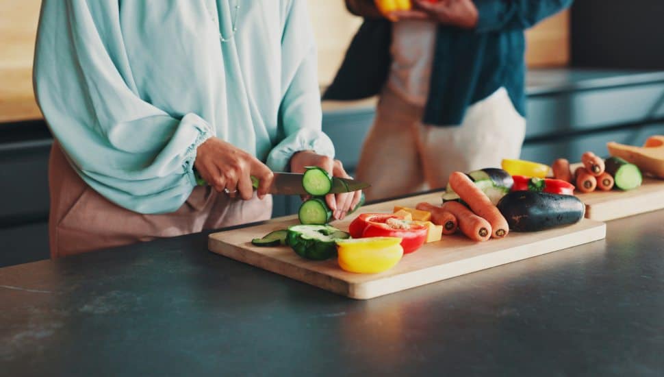 Hands, couple and cooking in kitchen with vegetables, knife and support with meal prep in home. People, organic ingredients and cutting board for food, diet and together for nutrition at apartment.