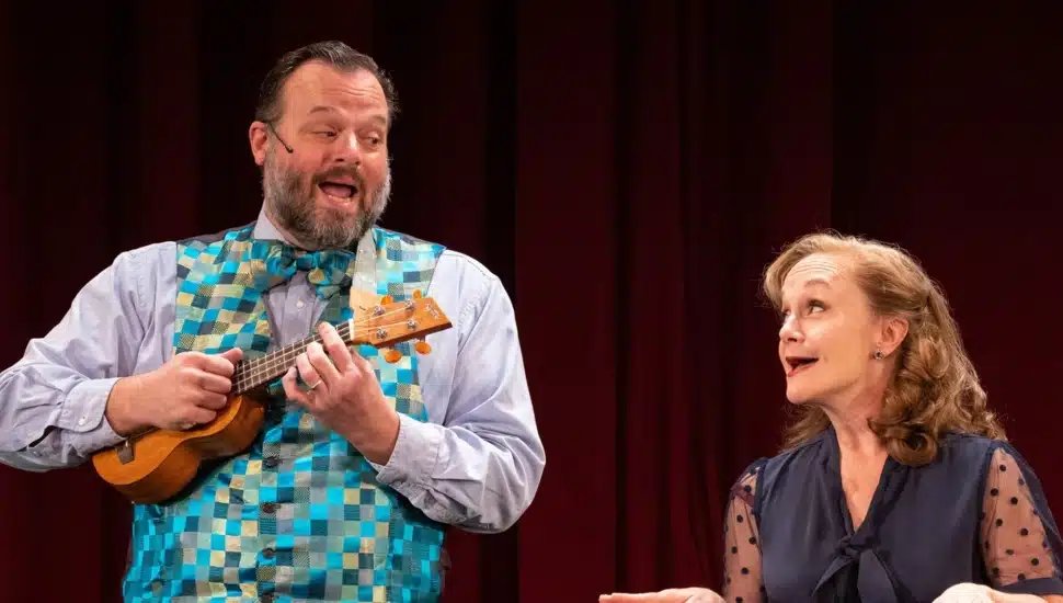 Scott Greer plays a ukulele in a colorful checkered vest and bow tie while Jennifer Childs looks on with delight during a performance of Two Outta Three at People's Light.