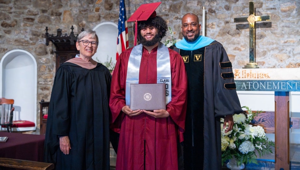 High school boy in cap and gown poses for picture school administrators at graduation.