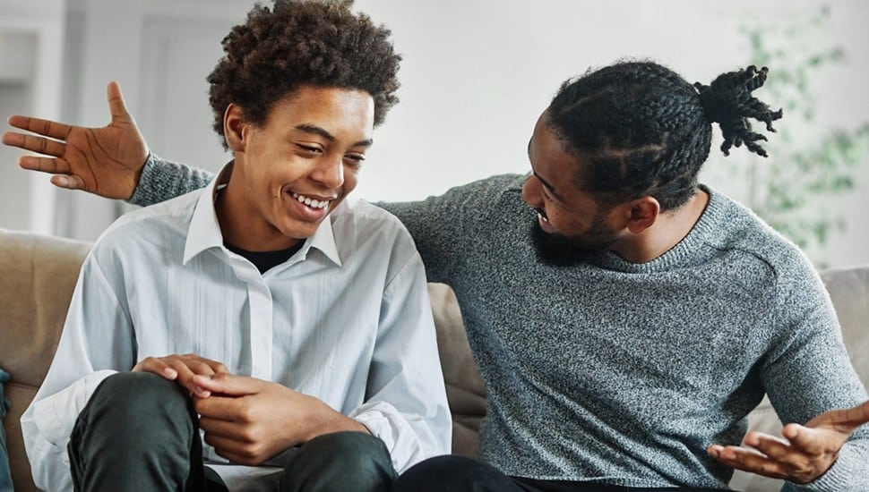 A father and son sit on the couch having a discussion, laughing.