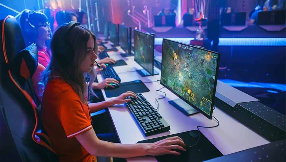 Female esports player wearing a gaming headset and sitting at a competitive gaming station, focused on a match while teammates and illuminated gaming equipment are visible in the background.