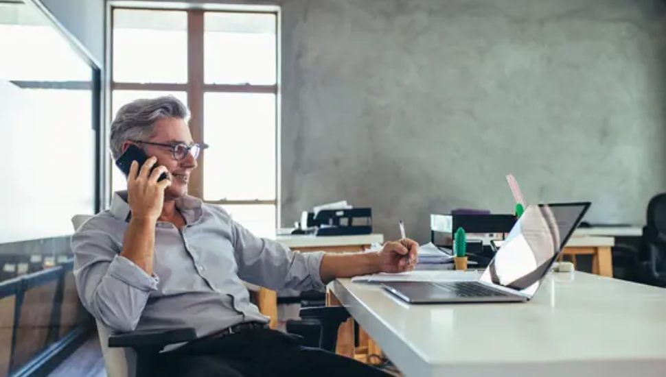 A small business owner smiles while talking on the phone and taking notes at his desk with a laptop open in front of him.